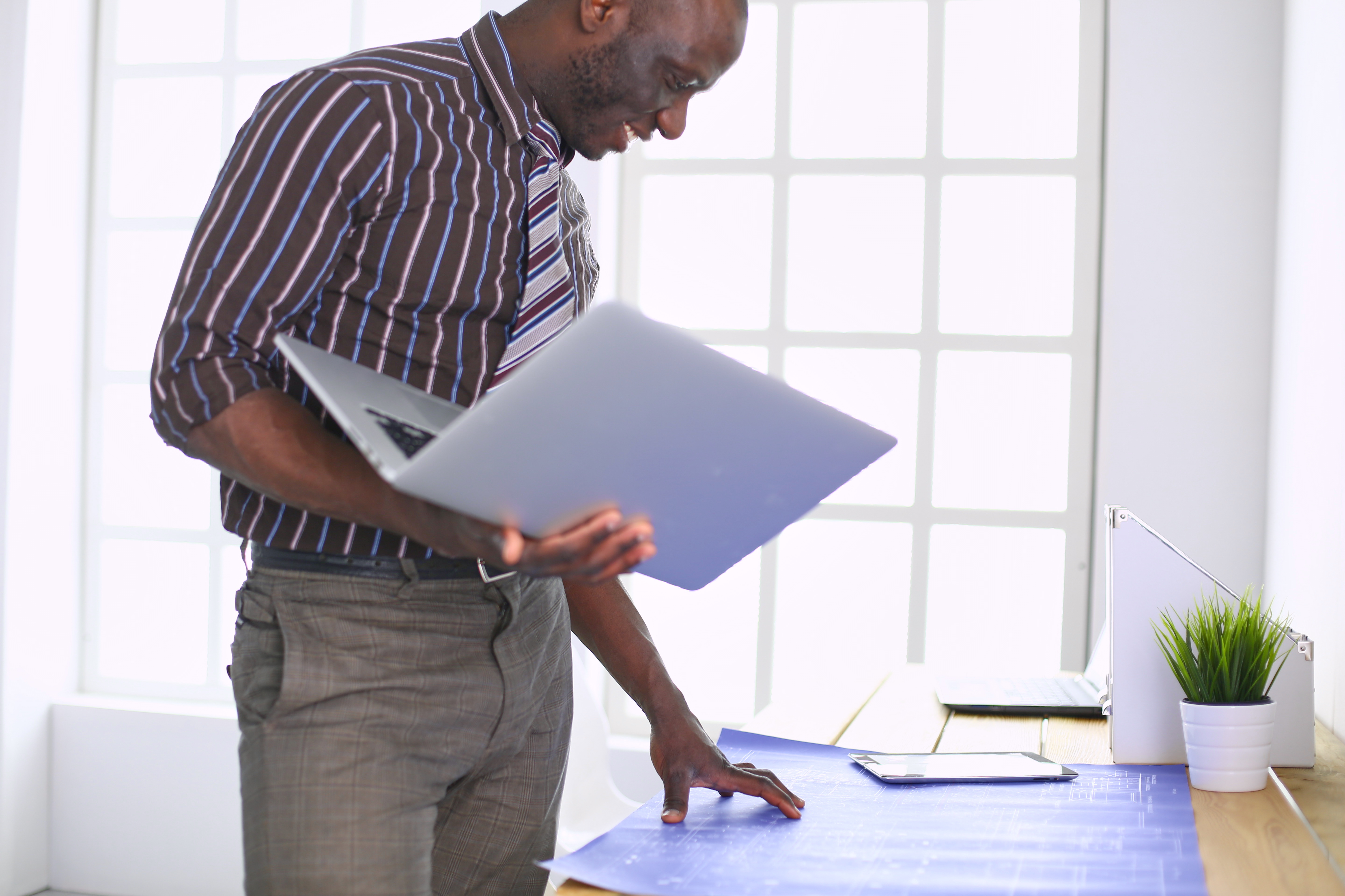 Black male drafter looking at blueprint holding laptop