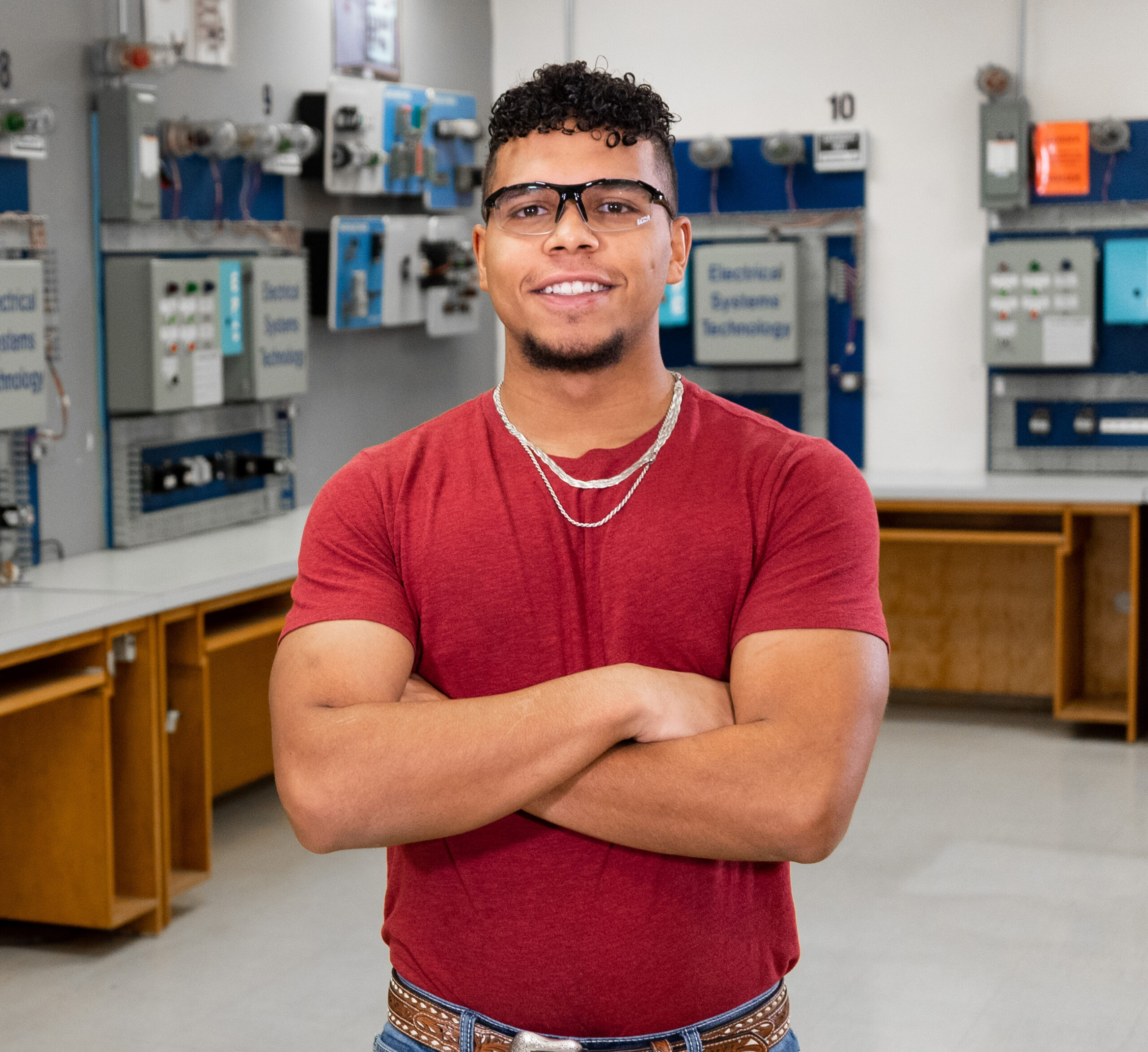 A young man in safety glasses stands in front of electrical powers training lab equipment with his arms crossed. He is smiling at the camera.