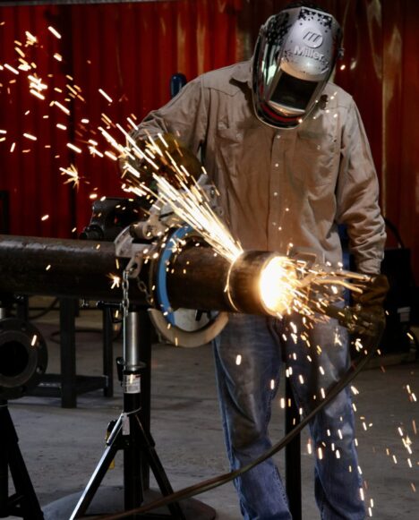 A welding student in a tan shirt and welding mask cuts a pipe to the appropriate length