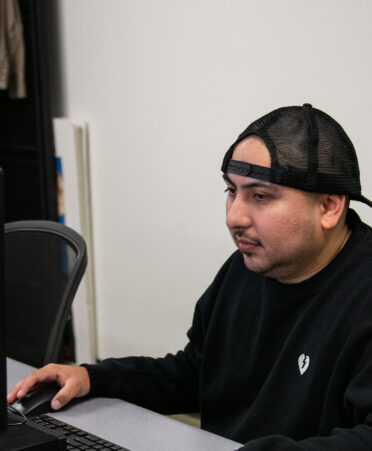 student wears black hat backwards with black sweatshirt and sits at a computer monitor