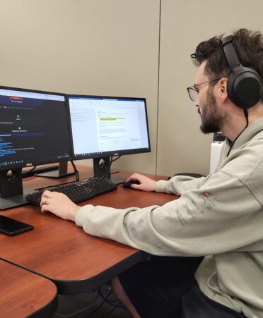 Grant Baird looks at computers while wearing a biege hoddie and black headphones