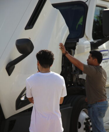 A student in a white shirt and orange shorts stands away from the camera looking at a white truck with its hood open. Another student wearing jeans and a green t-shirt looks at the truck's internals.