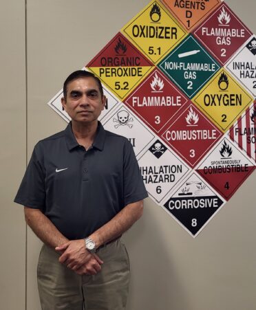 Hari stands wearing a black polo shirt against a wall with safety signs on it.