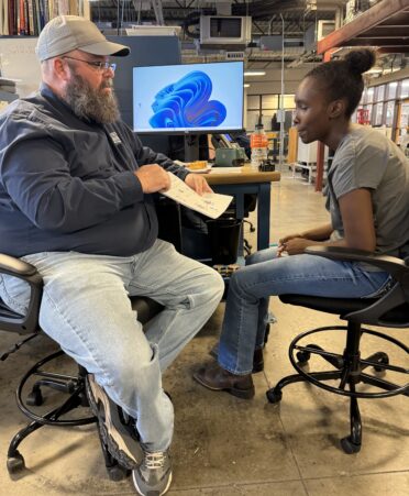 Jayme Palady is sitting on the left of the frame, pointing at the worksheet Amber Taylor completed to show her the problem he is explaining to her. Amber Taylor is sitting on the right and leaning toward the paperwork to see the diagrams.