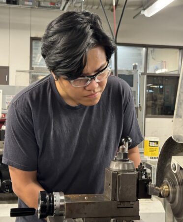 Andrew Doan wears safety glasses and a dark grey t-shirt while using a lathe to craft brass in a TSTC machine lab.