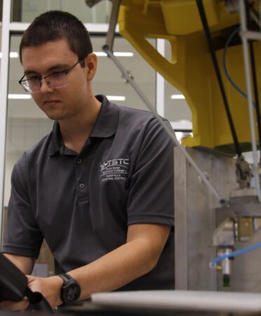 A Yellow robotic arm sits in the foreground. Andrey works with a black controller in the background while wearing a grey tstc shirt