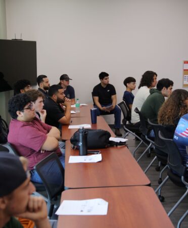 A group of students are sitting at desks. A backpack and various papers lay on the desks.