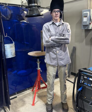 Michael Skufca stands in a welding bay with his arms crossed and his welding helmet on his head.