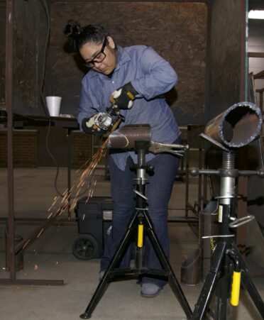 Paulina Maldonado uses a grinder on a metal pipe, causing sparks to fly toward the ground, to smooth edges to prepare to weld the pipe.