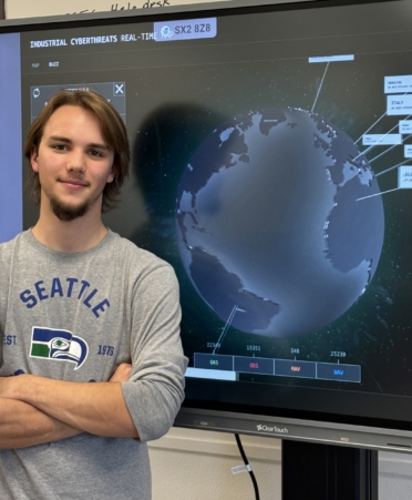 Blaise Deibert stands with arms crossed infront of a cyberthreat screen showing the globe with pop up notifications of threats in different countries.