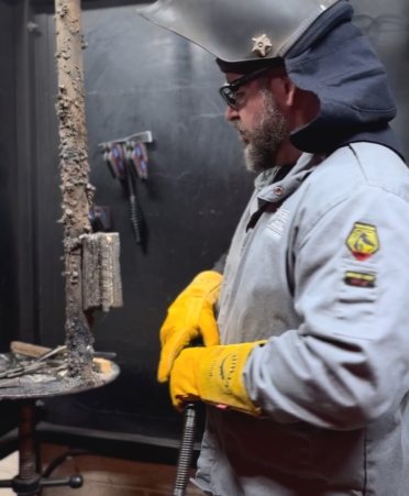 Brad Panneton stands with a welding helmet on his head in a welding booth at TSTC looking at a metal plate he TIG welded.