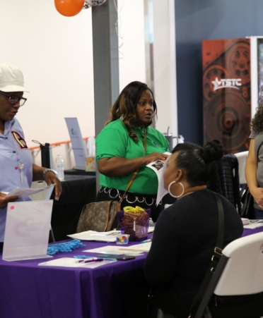 People wearing a blue shirt and green shirt respectively stand across from a resource booth with two representatives on the other side, wearing black and gray shirts respectively.