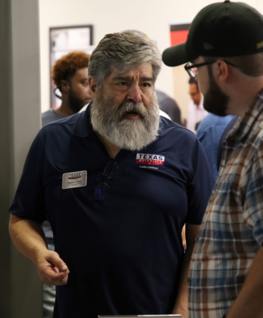 A man with a gray beard and wearing a Texas Materials blue shirt meets with a student wearing a plaid shirt and ballcap.