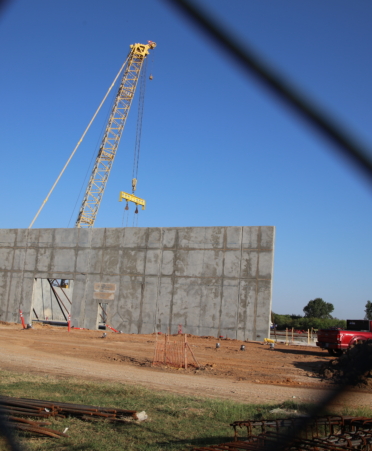 A yellow crane rests behind the gray tilt walls. A chain link fence frames the two.