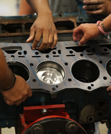 People work with a diesel engine part that is silver with blue stripes running down it. Only the hands of the people are seen.