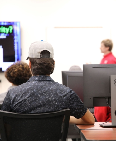 A high school students listens to a presentation. He is wearing a gray ball cap and blue speckled shirt. Computers sit on a desk next to him.