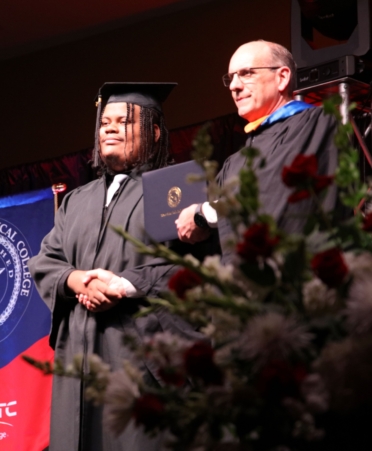 A graduate in cap and gown shakes hands with an instructor while receiving his diploma. Flowers sit in the foreground.