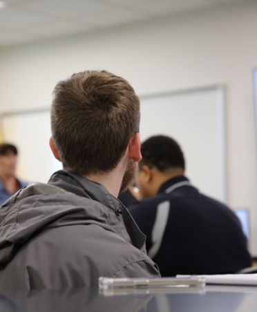 A student with a gray jacket on listens to a lecture with his back turned away from the camera.