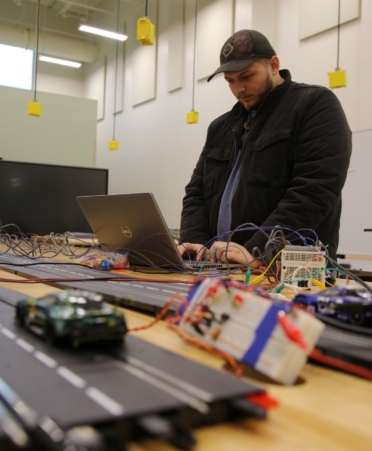 A miniature racetrack sits on a table with Charles wearing black coat working on a laptop sits in the background.