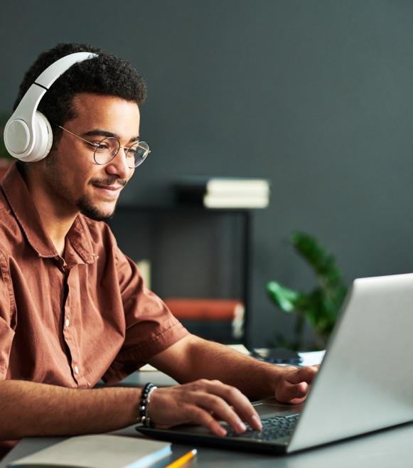 Young smiling man in headphones typing on laptop keyboard while sitting by workplace and taking part in online webinar or lesson