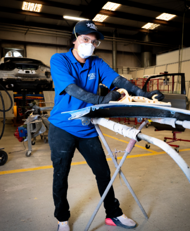 A female student wearing a blue tstc shirt and a white mask works on a car part.